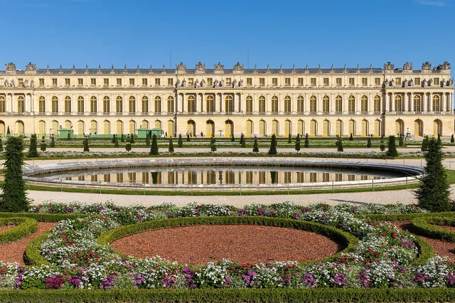The long golden facade of the Palace of Versailles behind a round pond and colourful flowerbeds in the Parterre du Midi