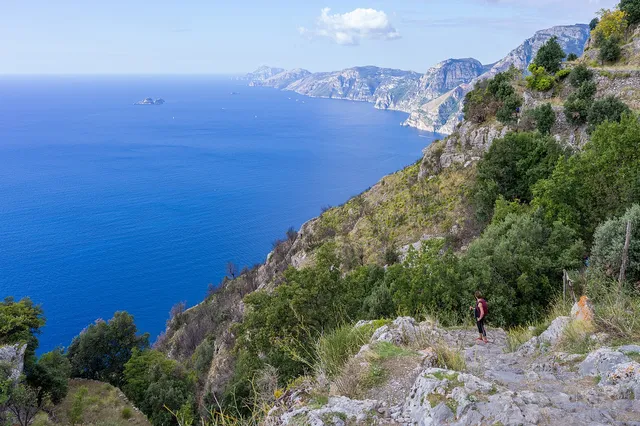A rocky hiking path along green cliffs high above the deep blue sea of the Amalfi Coast, with headlands stretching into the distance