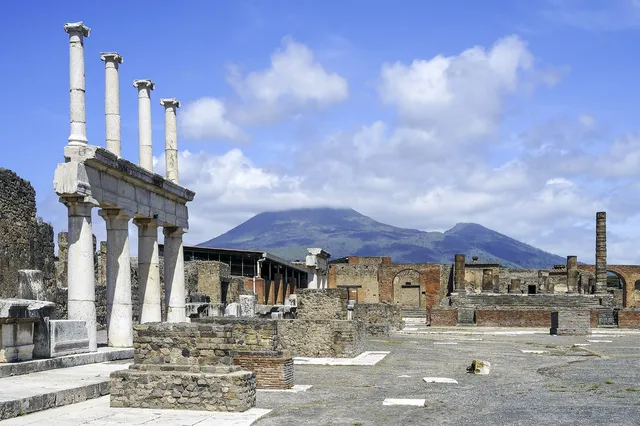 White stone columns of the Forum in Pompeii with the slopes of Mount Vesuvius rising behind under a blue sky