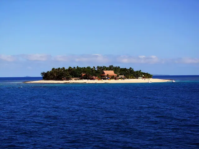 A small palm-covered coral island with a white sand beach and thatched huts in the Mamanuca Islands, surrounded by deep blue sea