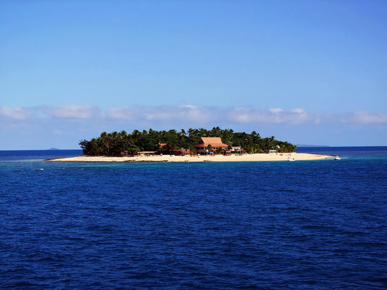 A small palm-covered coral island with a white sand beach and thatched huts in the Mamanuca Islands, surrounded by deep blue sea