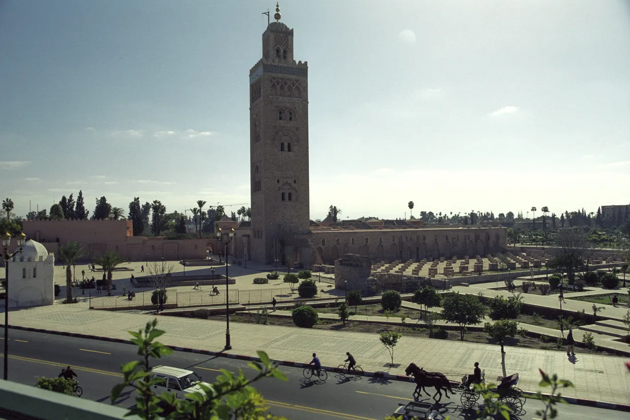 The minaret of the Koutoubia Mosque in Marrakech rising above palm trees and gardens, with a horse-drawn calèche on the road in front