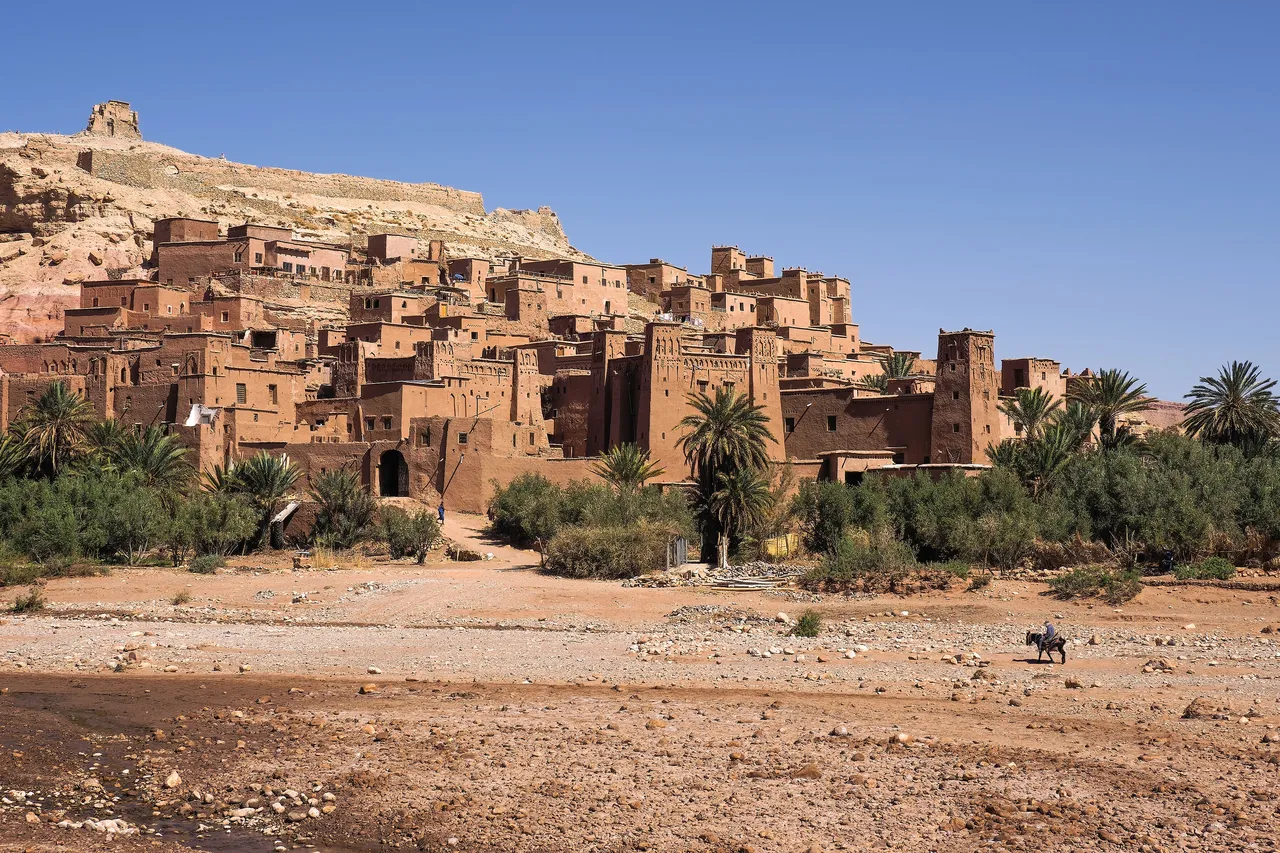 The earthen kasbahs of the ksar of Aït Benhaddou rising above palm trees and a dry riverbed in southern Morocco