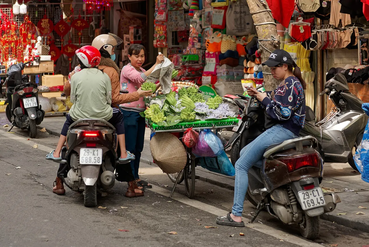 A street vendor selling greens from a bicycle to people on motorbikes in front of shops in Hanoi's Old Quarter